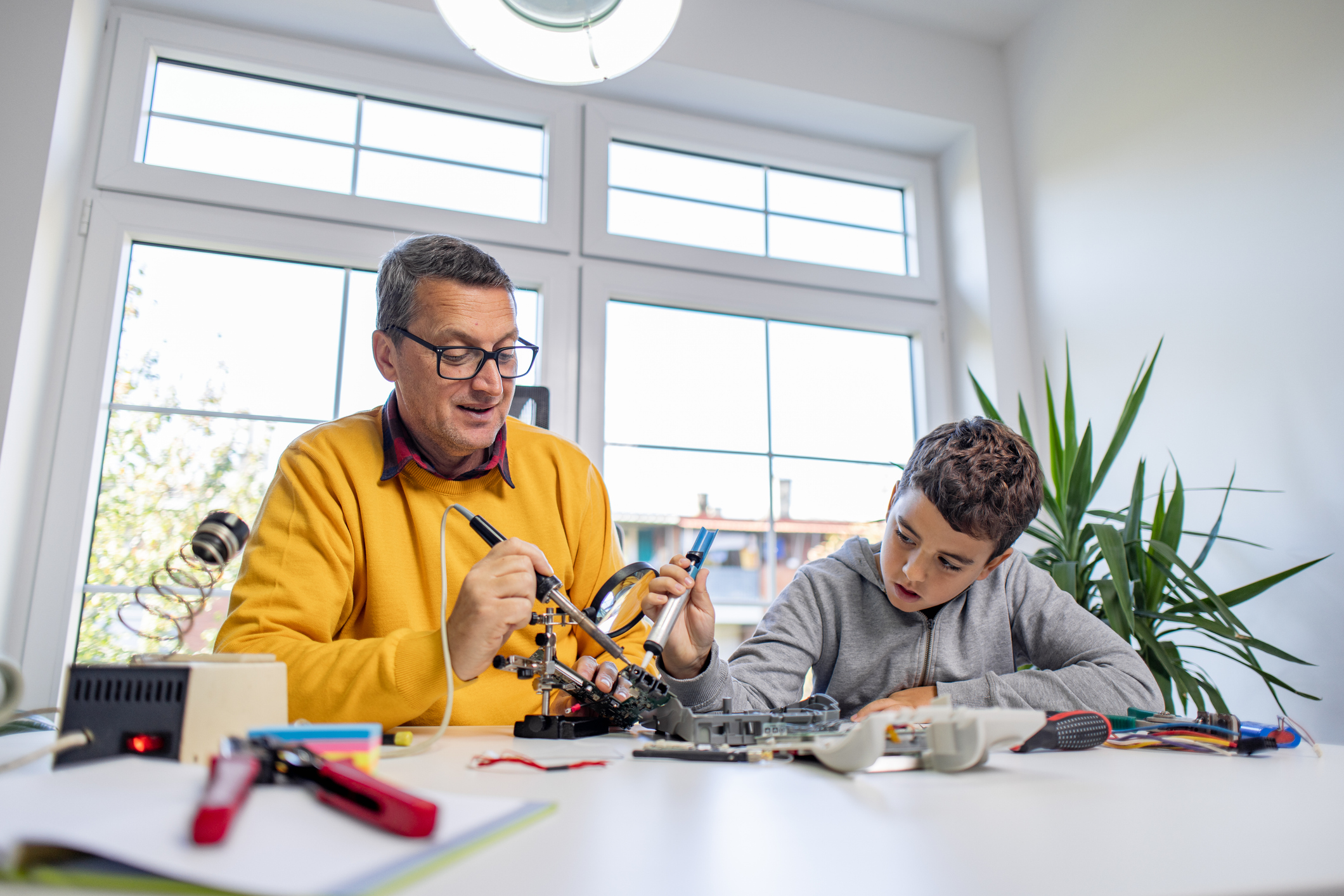 An adult and a child sit at a table working on an electronics project, using tools and components in a bright room with large windows.