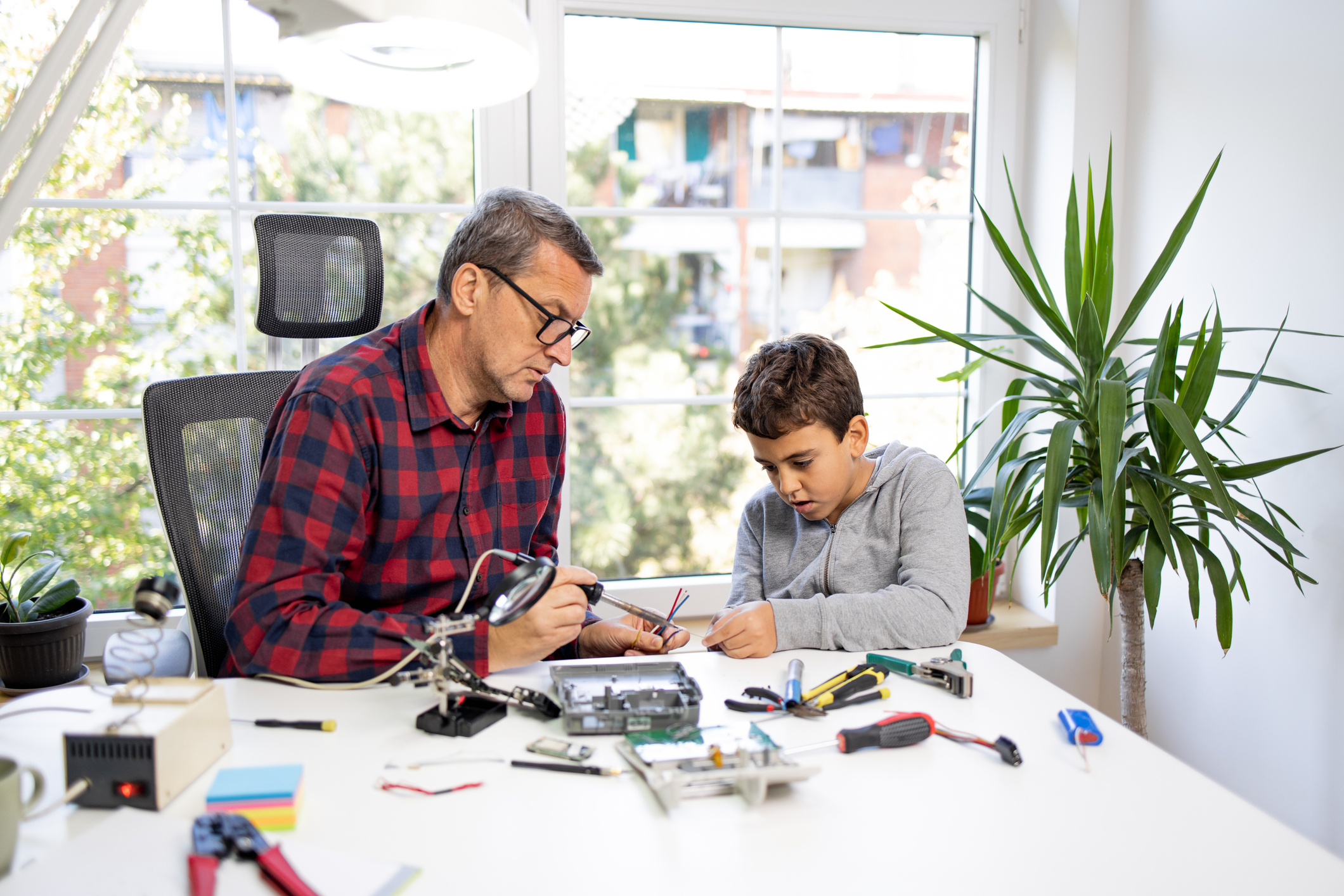 An adult and a child sit at a desk assembling electronic components, with tools and parts spread out around them in a bright room with large windows and plants.