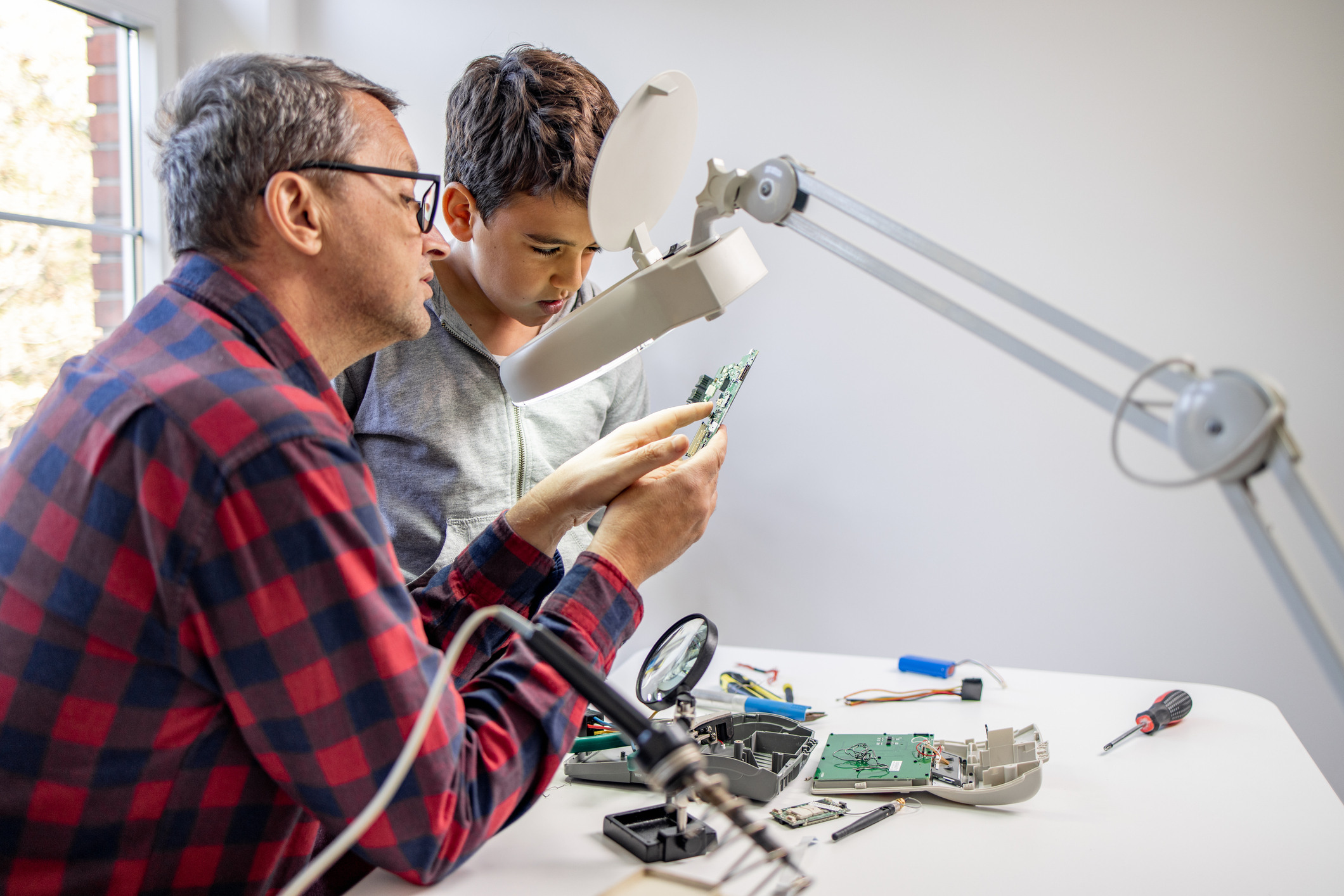 An adult and a child examine electronic components at a workbench, using a magnifying lamp and surrounded by disassembled device parts and tools.