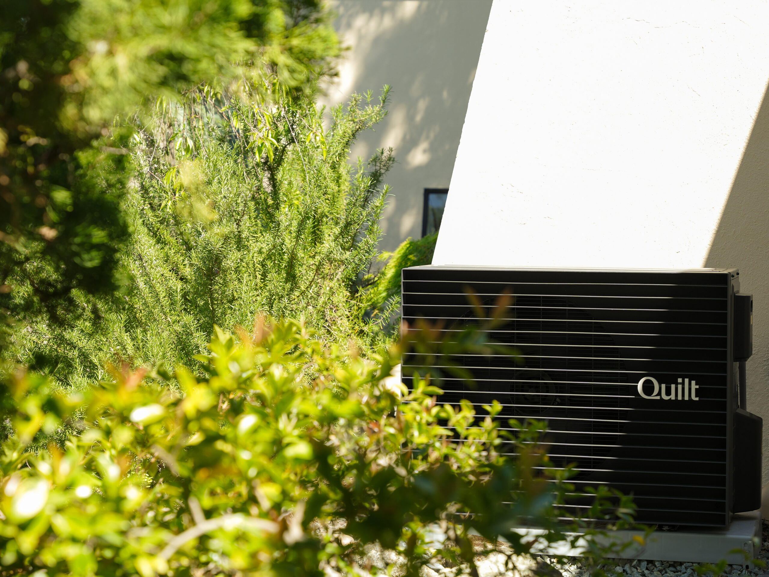 A black outdoor AC unit labeled "Quilt" is mounted on a light-colored wall, partially surrounded by green foliage and plants.
