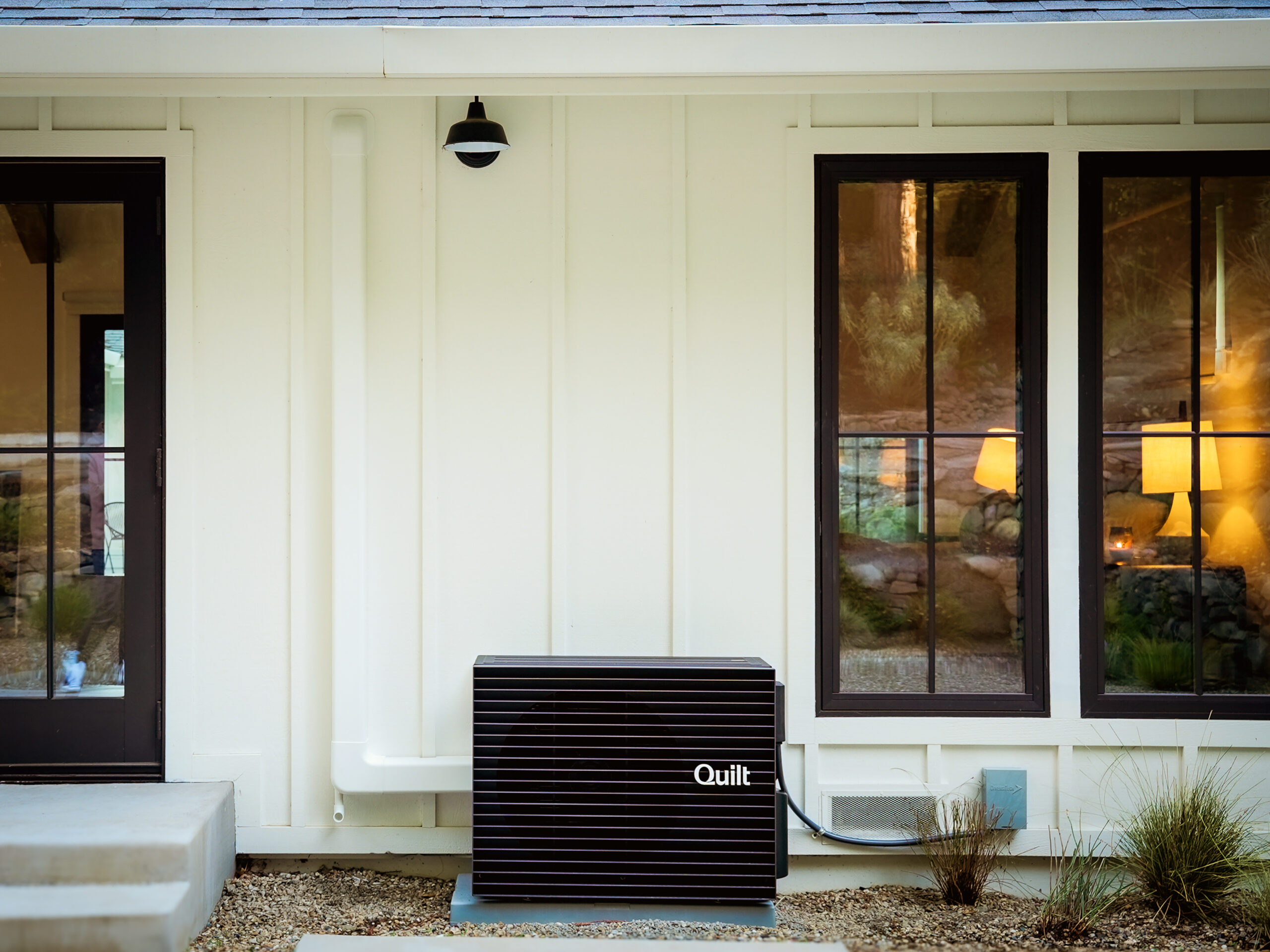 A black Quilt heat pump unit is installed outside a white house, below two large windows and next to a glass door.