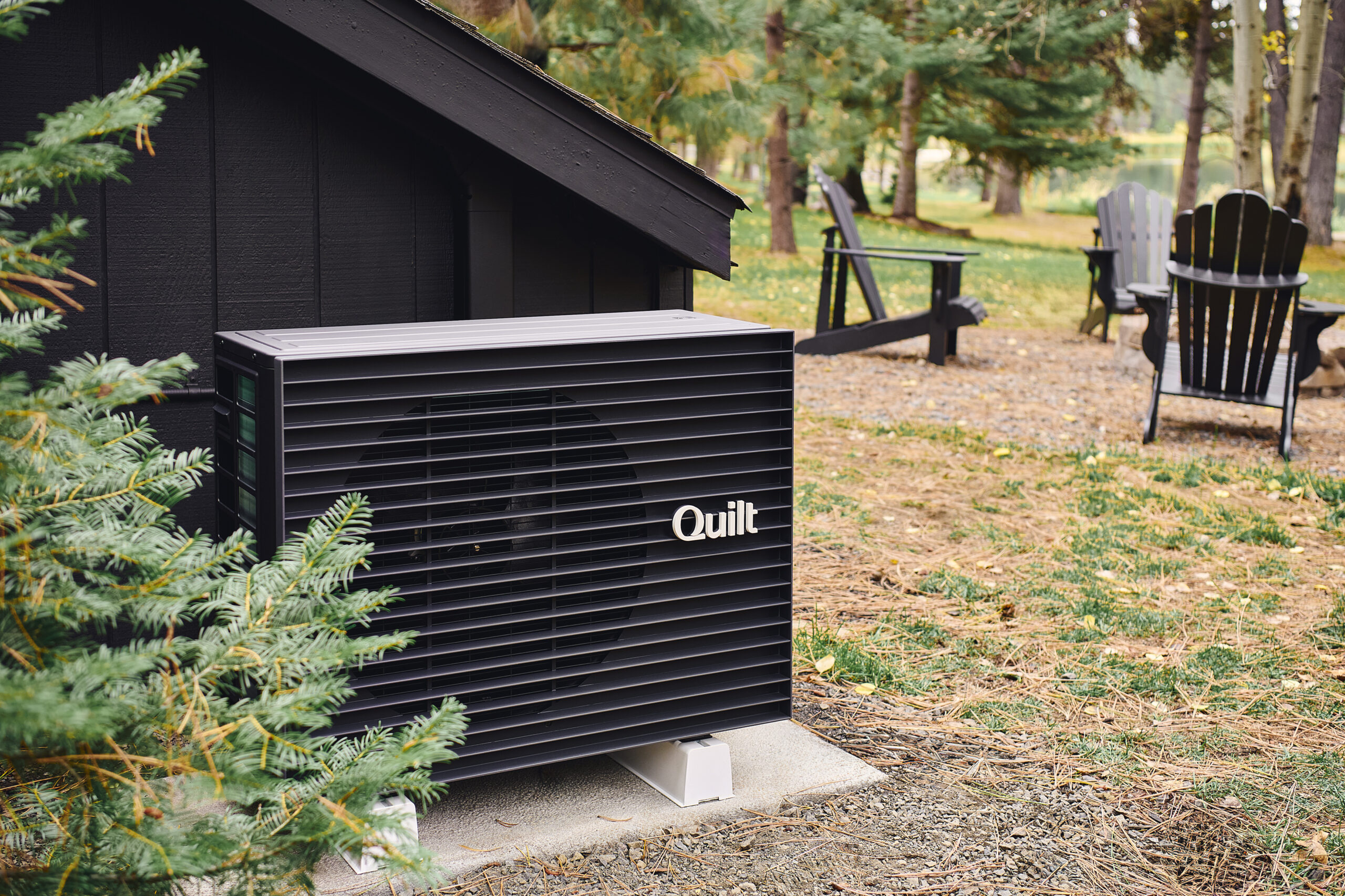 A Quilt heat pump unit is installed outside a dark wooden building near evergreen trees, with outdoor chairs and a natural landscape in the background.