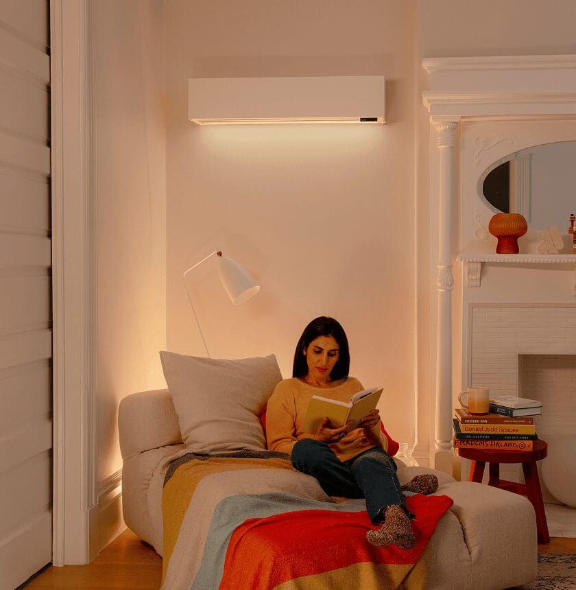 A woman sits on a cushioned chair reading a book under a floor lamp in a cozy, well-lit living room with a colorful blanket and a wall-mounted air conditioner.