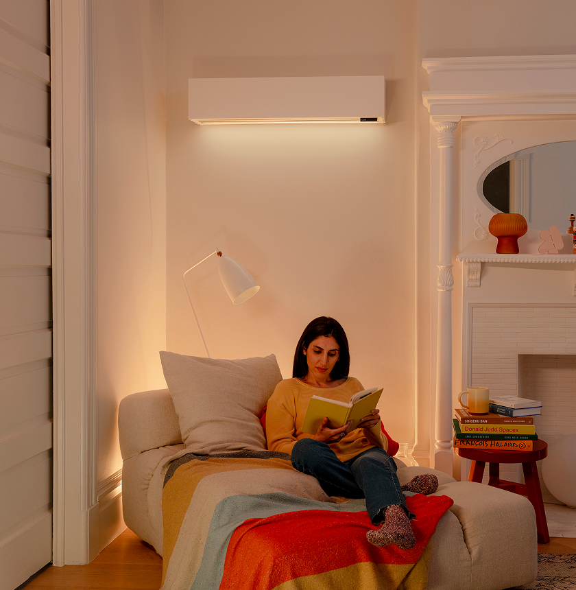A woman sits on a cushioned chair reading a book under a floor lamp in a cozy, well-lit living room with a colorful blanket and a wall-mounted air conditioner.
