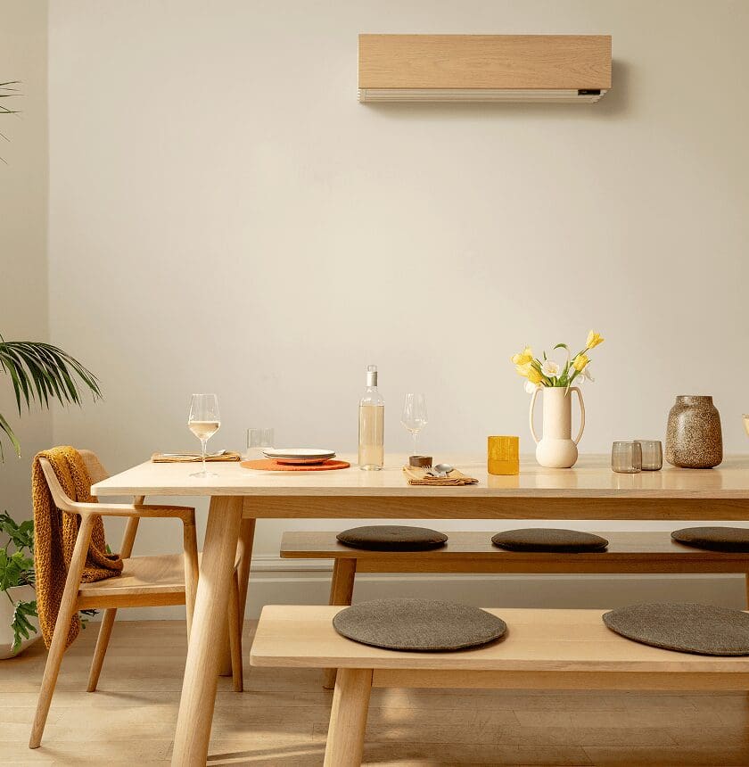 A modern dining area with a wooden table, bench seating with round cushions, wine glasses, plates, a bottle, a jug with yellow flowers, and neutral decor.