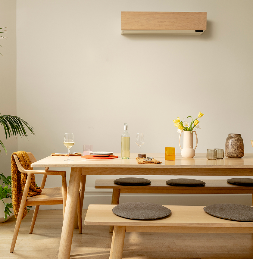 A modern dining area with a wooden table, bench seating with round cushions, wine glasses, plates, a bottle, a jug with yellow flowers, and neutral decor.