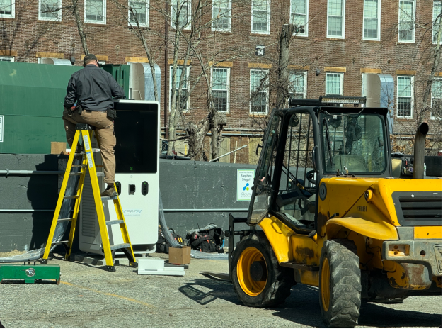 A man stands on a yellow ladder working on an electrical unit near a green dumpster, with a yellow construction vehicle parked nearby in a lot.