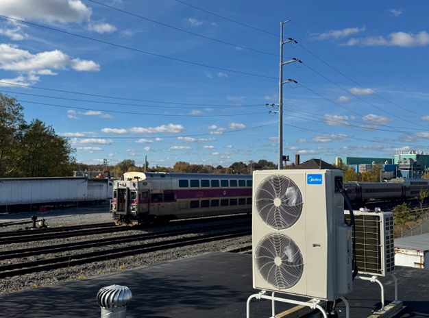 A commuter train travels on railway tracks under power lines, with an air conditioning unit installed on a nearby rooftop in the foreground.