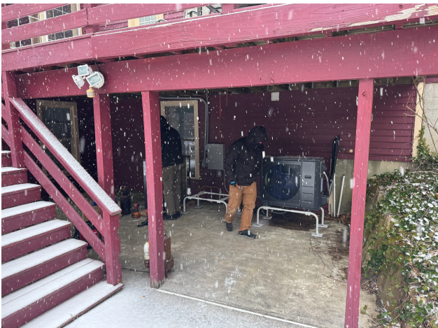 Two people stand under a deck next to a heat pump unit during snowfall, with snow lightly accumulating on the ground and stairs.