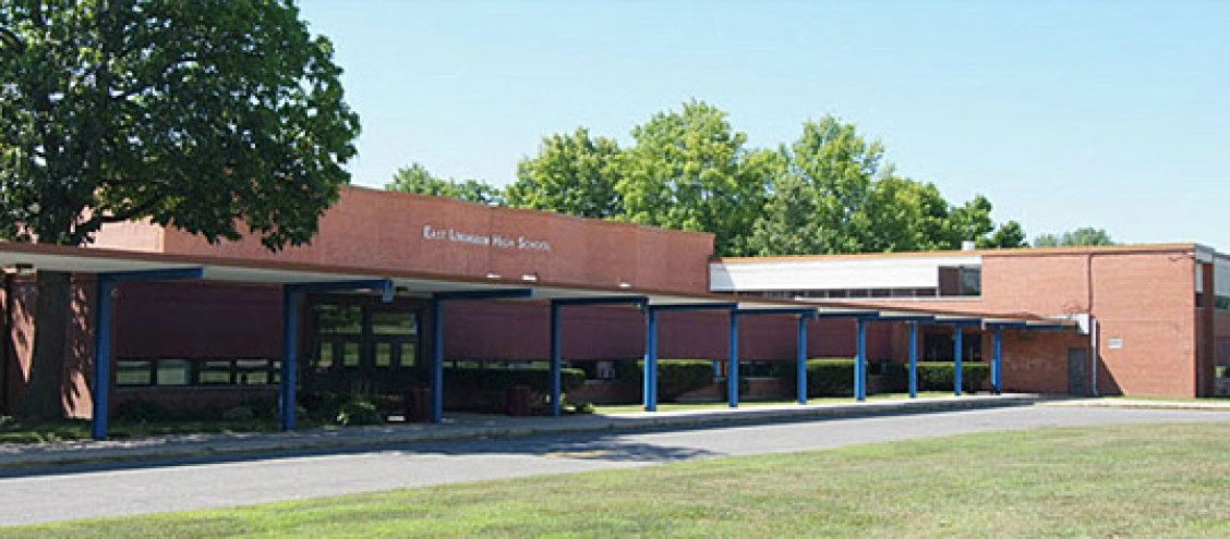 A single-story red-brick school building with blue pillars, large windows, and a flat roof, surrounded by trees and grass. The sign reads "East Arkansas High School.