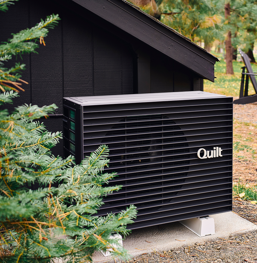 A Quilt-branded heat pump unit is installed outside a dark-painted building, partially obscured by evergreen branches.