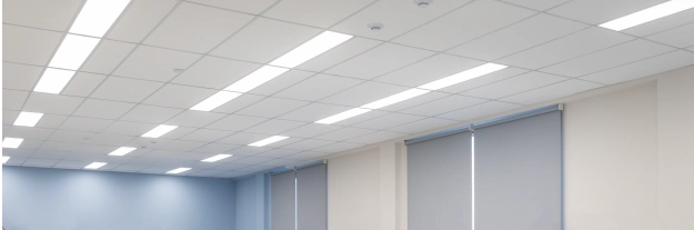 A room with a white suspended ceiling, rectangular LED light panels, and closed gray roller blinds on the windows.