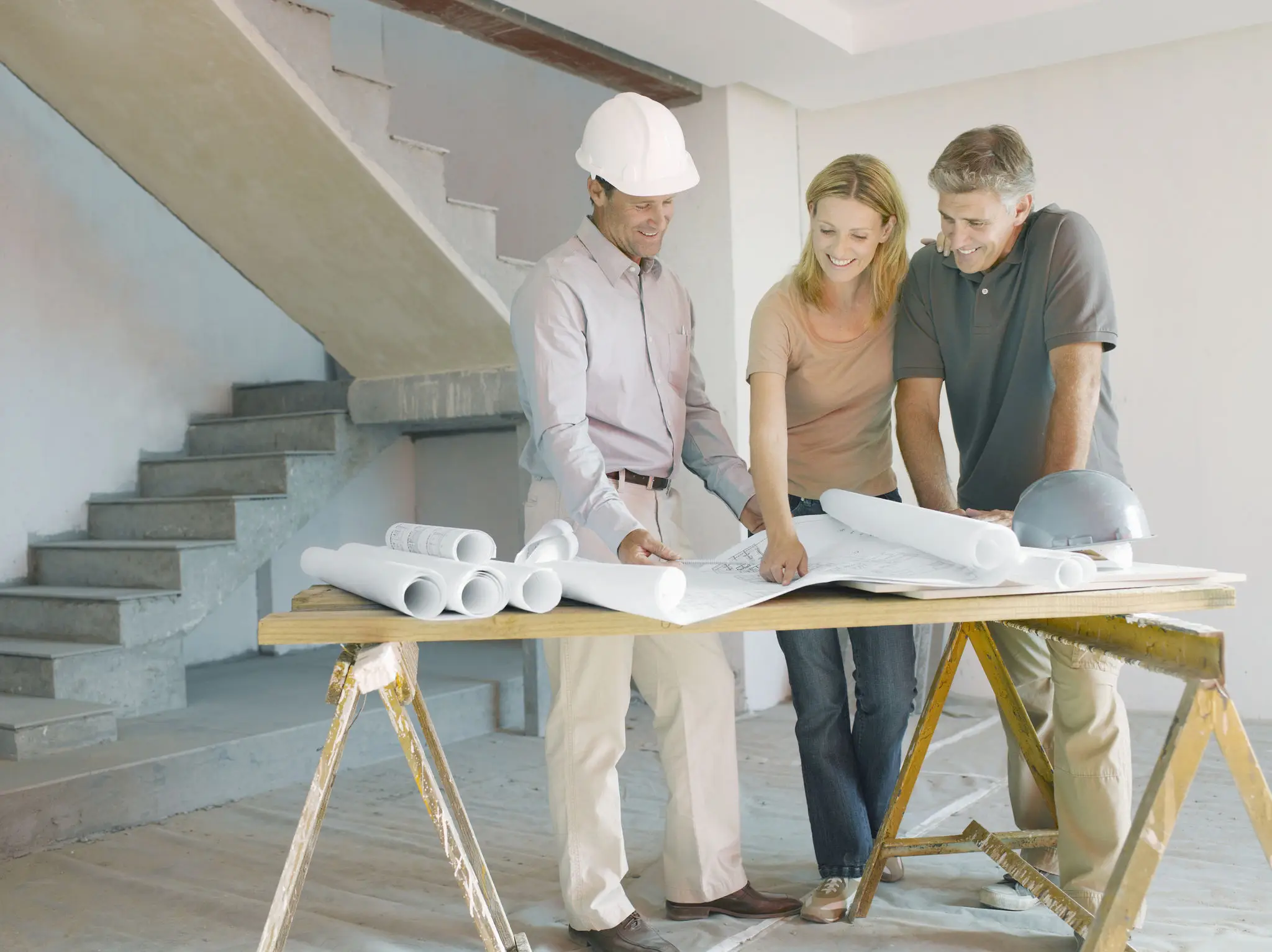 Three people review blueprints on a table at a construction site. One wears a hard hat. Stairs and construction materials are visible in the background.