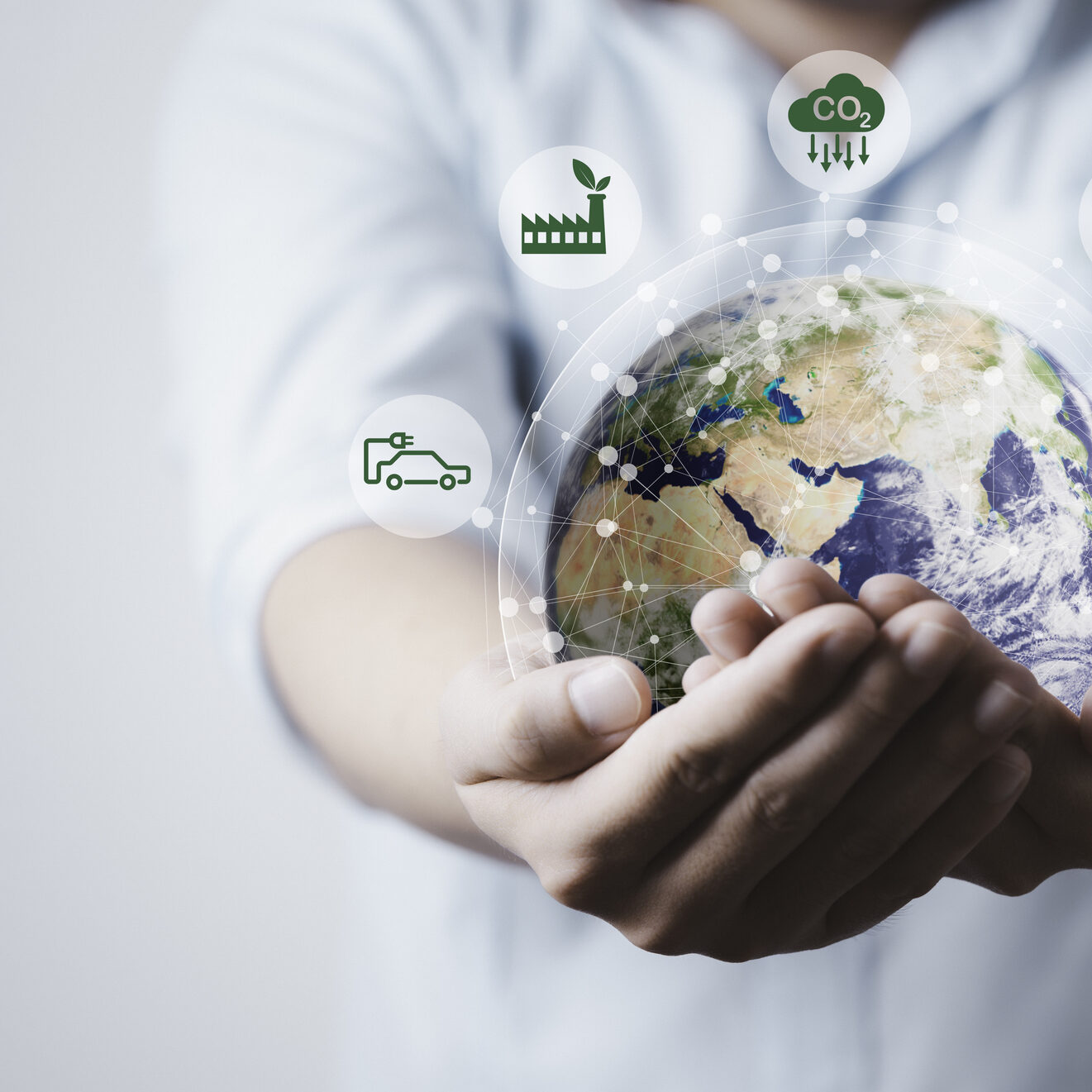 A person holds a digital globe with sustainable energy and environmental icon graphics, including wind, solar, electric car, and factory symbols.