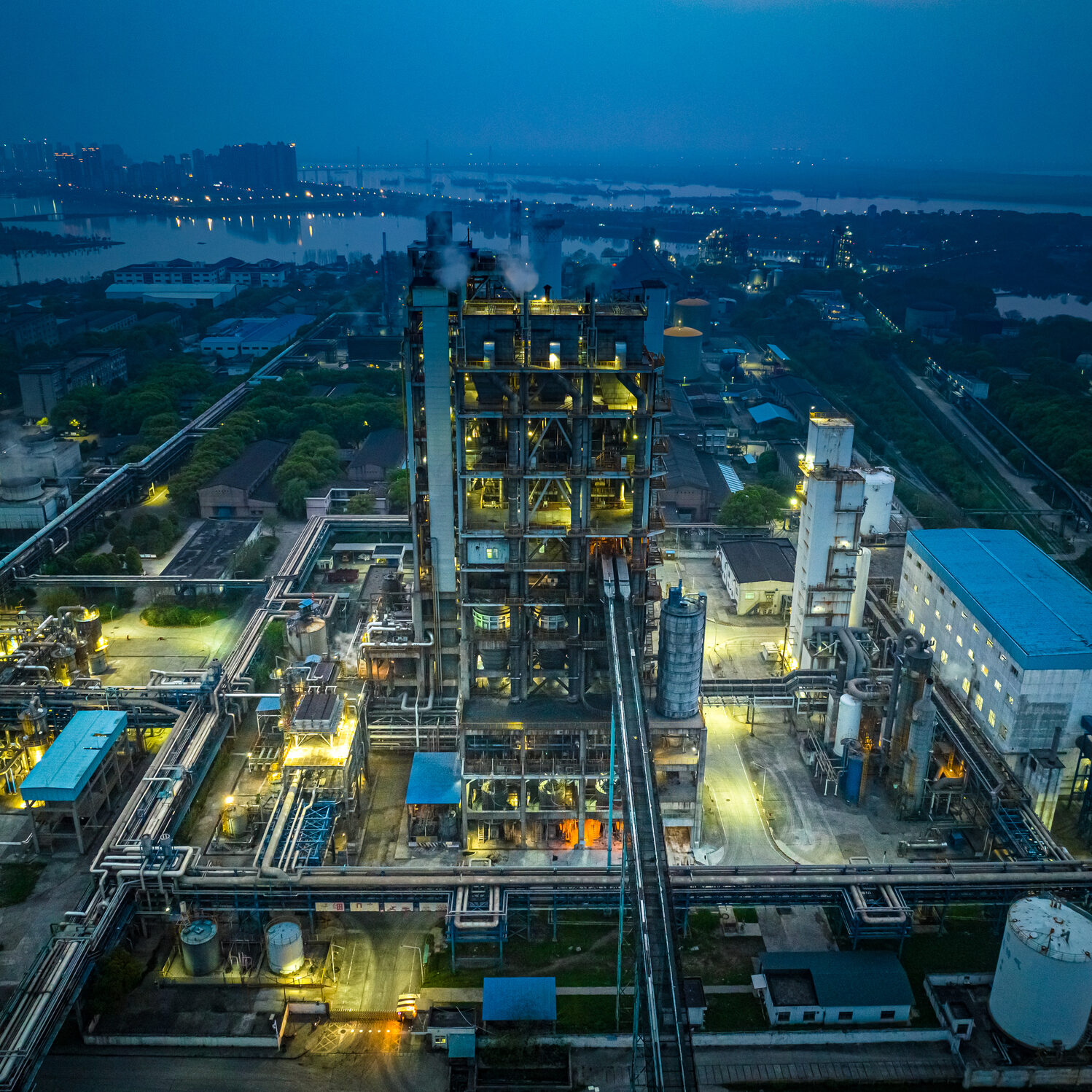 Aerial view of an industrial plant illuminated at dusk, with smokestacks, storage tanks, and various buildings surrounded by pipelines and a river in the background.